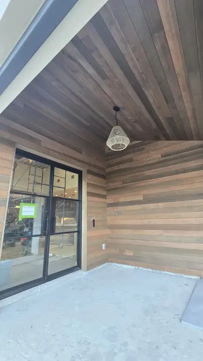 Covered exterior room wrapped in horizontal wood cladding with a matching wood ceiling and black-framed door.
