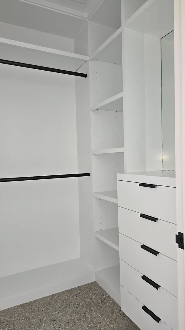 White closet cabinetry with adjustable shelves, black hanging rails, drawers with black pulls and a mirror.